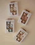 Four boxes of FRIGG Baby's First Pacifier Floral Heart (Blush) 4-Pack, made in Denmark, are arranged on a light surface. Each box displays two pastel pacifiers through a clear window, highlighting the charming floral heart design.