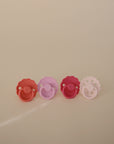 Four FRIGG Natural Rubber Pacifiers from the FRIGG 4-Pack are lined up on a light surface, featuring red, pink, and purple shades with unique edge designs on each Danish pacifier.
