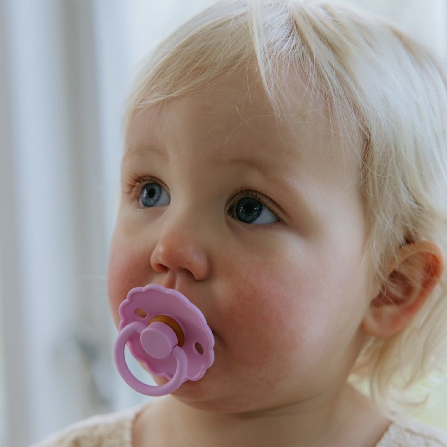 A young toddler with light blonde hair and blue eyes looks up while using a pink FRIGG Natural Rubber Pacifier from the FRIGG 4-Pack. Soft natural light illuminates their face, and a blurred indoor background is visible.