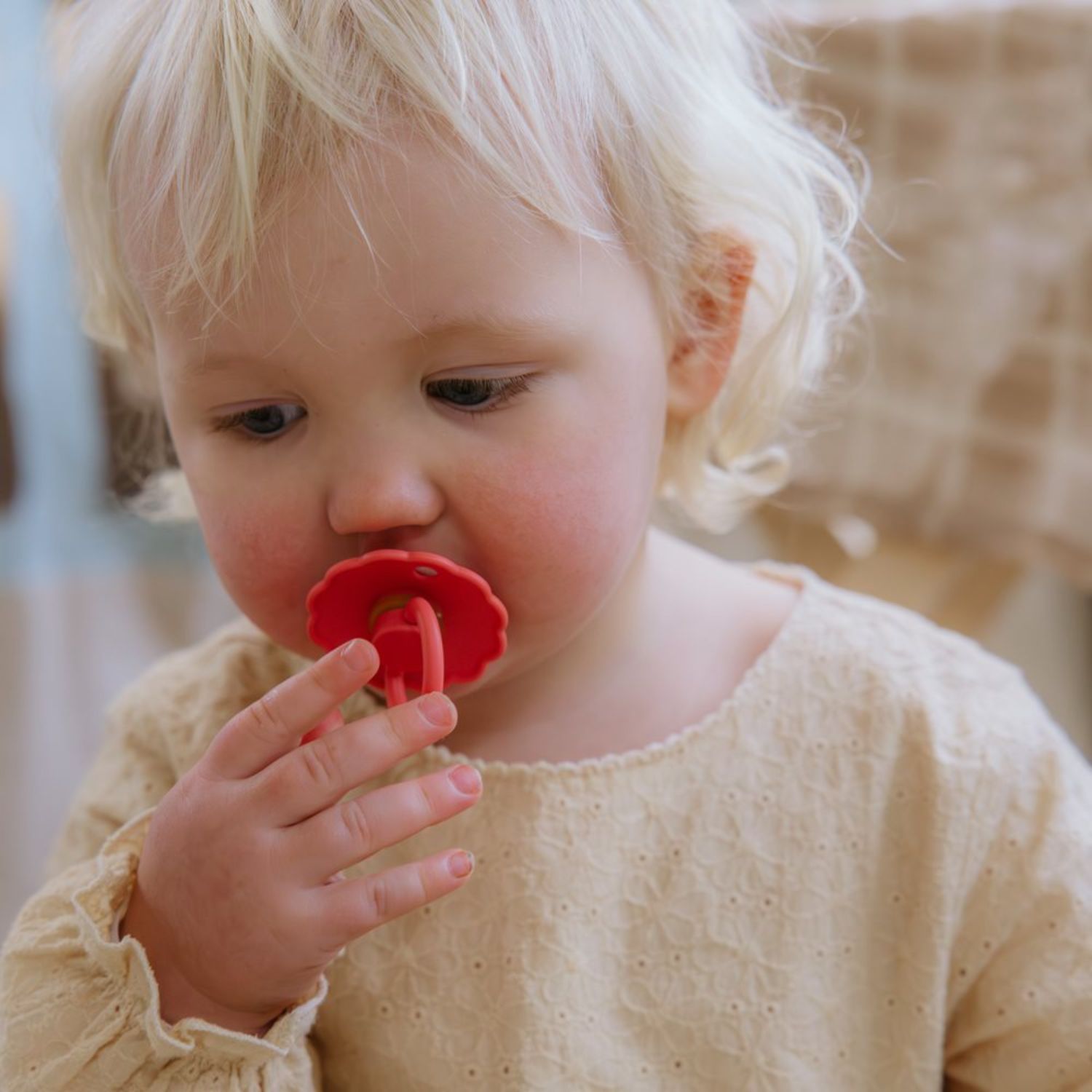 A young child with blonde hair thoughtfully looks down while holding a FRIGG Natural Rubber Pacifier from the FRIGG 4-Pack in their mouth, dressed in a light textured top against a soft, blurred background.