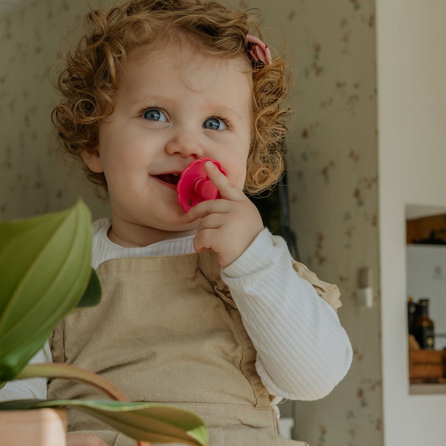 A smiling young child with curly hair and blue eyes holds a pink pacifier from the FRIGG Natural Rubber Pacifier 4-Pack, dressed in beige over a white shirt, standing indoors near a green plant and floral wallpaper.