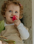 A smiling young child with curly hair and blue eyes holds a pink pacifier from the FRIGG Natural Rubber Pacifier 4-Pack, dressed in beige over a white shirt, standing indoors near a green plant and floral wallpaper.