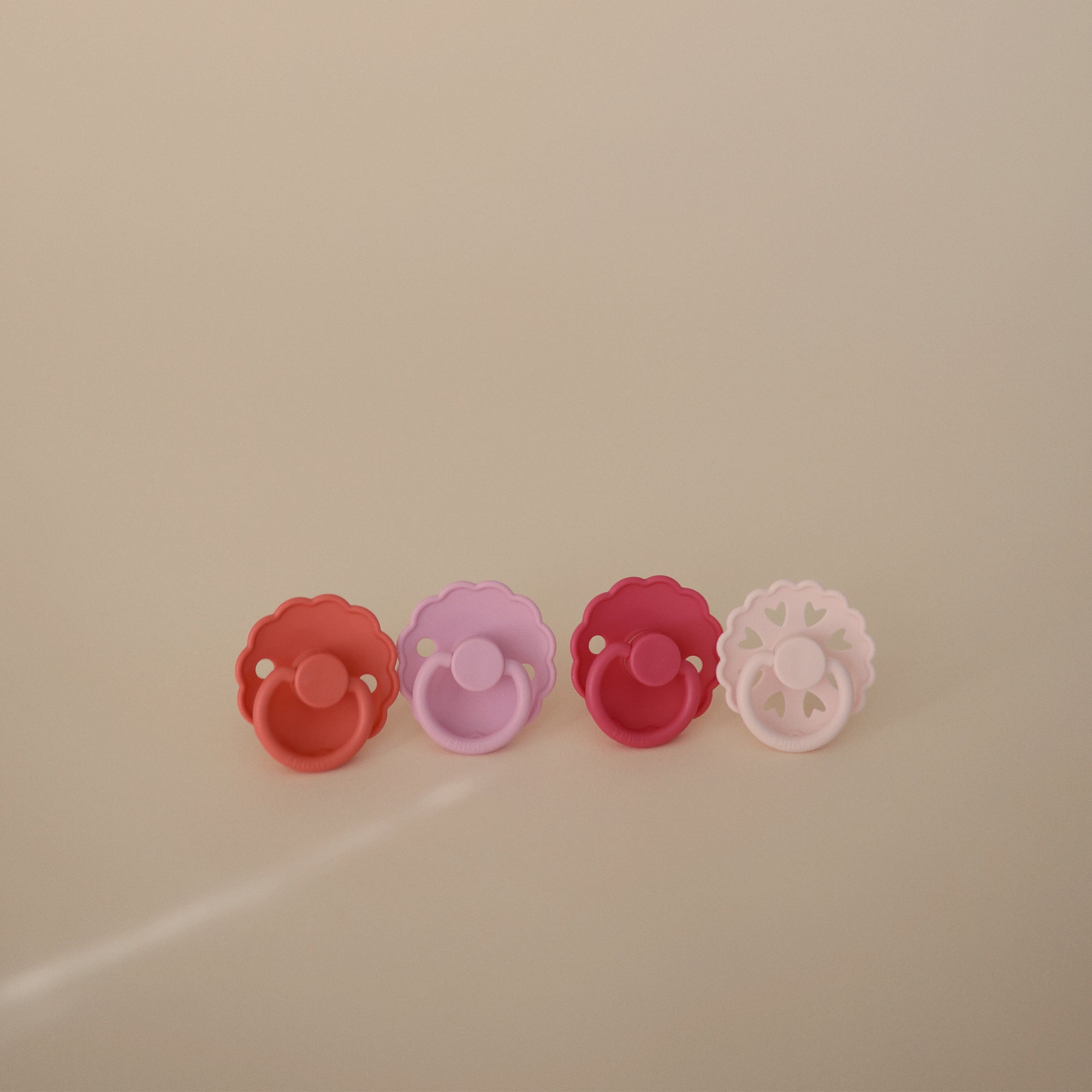 Four FRIGG Natural Rubber Pacifier 4-Pack pacifiers—red, purple, pink, and white—are displayed in a row with heart-shaped cutouts. Made by FRIGG from natural rubber, they offer soothing comfort with a stylish Danish design.