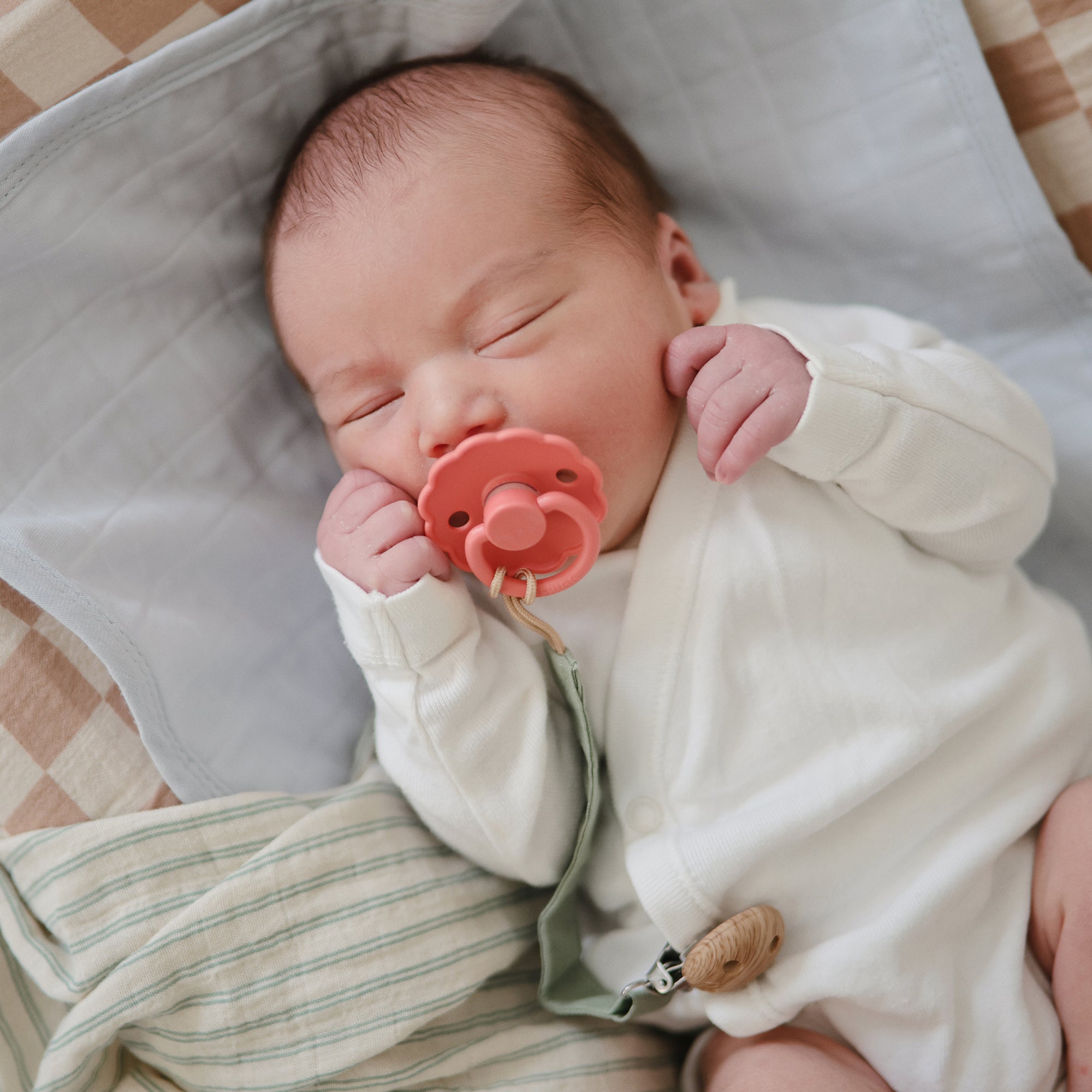 A newborn in a white onesie sleeps peacefully on a soft blanket, using a FRIGG Natural Rubber Pacifier from the FRIGG 4-Pack. Their eyes are closed, and the pacifier is gently held in their mouth.