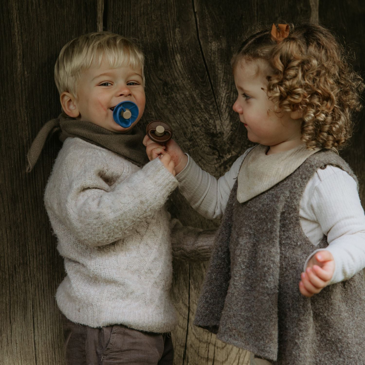 A young boy with a blue FRIGG Natural Rubber Pacifier and a girl holding a brown Danish pacifier stand together, smiling in cozy, neutral outfits against a rustic wooden backdrop. Both pacifiers are from the FRIGG Natural Rubber Pacifier 4-Pack.