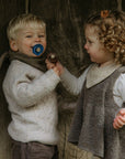 A young boy with a blue FRIGG Natural Rubber Pacifier and a girl holding a brown Danish pacifier stand together, smiling in cozy, neutral outfits against a rustic wooden backdrop. Both pacifiers are from the FRIGG Natural Rubber Pacifier 4-Pack.