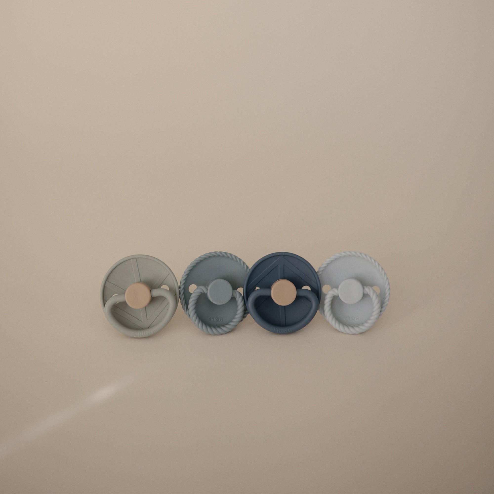 The FRIGG Natural Rubber Pacifier 4-Pack by FRIGG features four round pacifiers in muted gray, blue, and navy tones, neatly arranged against a neutral beige background.