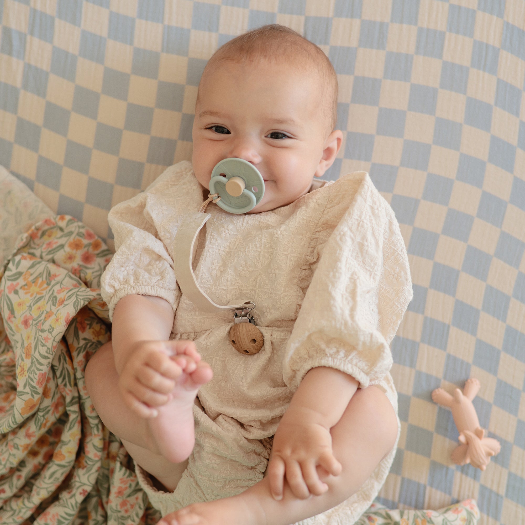 A smiling baby in a cream outfit lies on a blue and white checkered blanket, holding their foot and sucking on a FRIGG Natural Rubber Pacifier 4-Pack by FRIGG. Nearby are a floral blanket and a small toy.