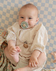A smiling baby in a cream outfit lies on a blue and white checkered blanket, holding their foot and sucking on a FRIGG Natural Rubber Pacifier 4-Pack by FRIGG. Nearby are a floral blanket and a small toy.