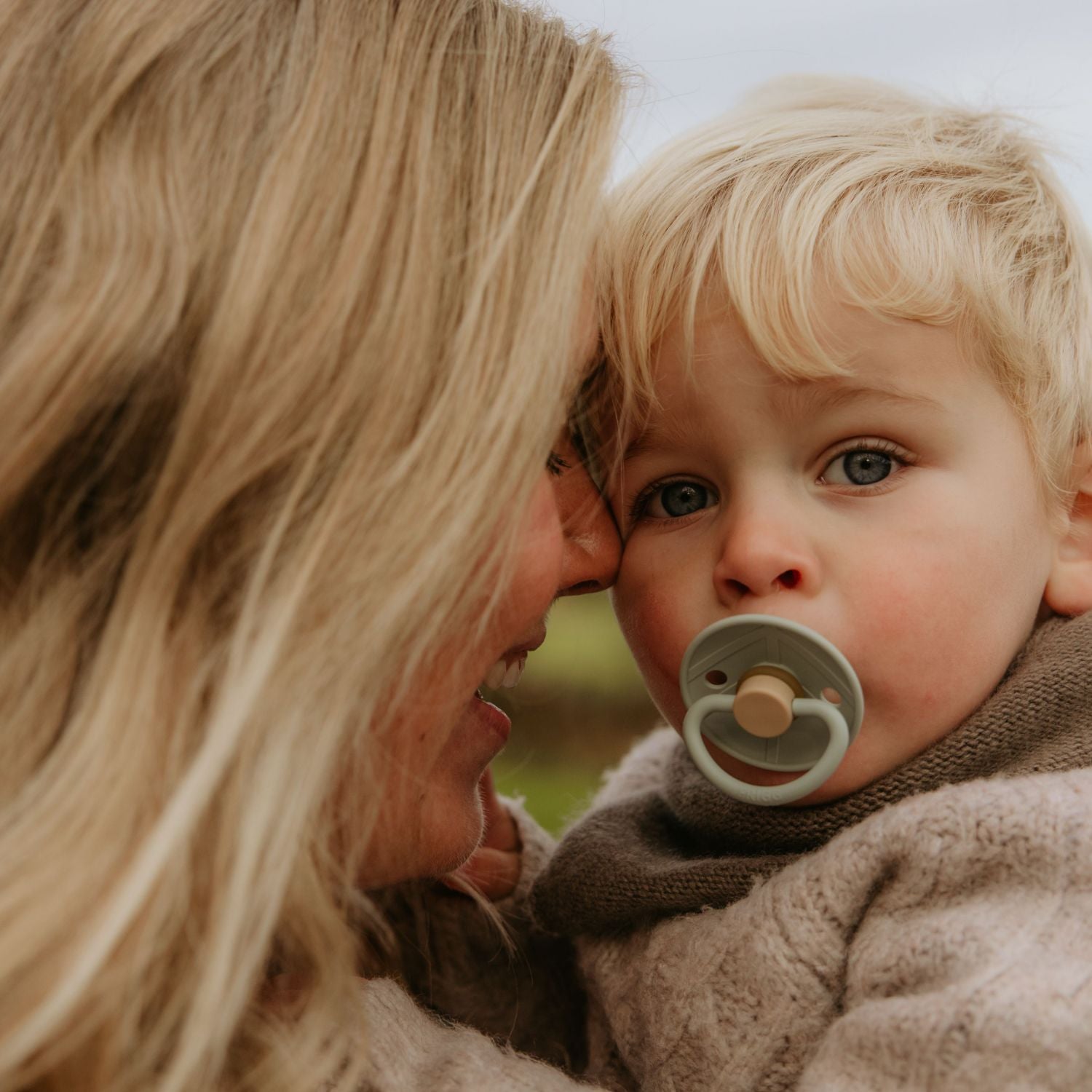 A woman with long blonde hair lovingly holds a blue-eyed child sucking on a FRIGG Natural Rubber Pacifier. Both are warmly dressed, and the pacifier is from the FRIGG 4-Pack collection.