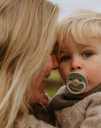 A woman with long blonde hair lovingly holds a blue-eyed child sucking on a FRIGG Natural Rubber Pacifier. Both are warmly dressed, and the pacifier is from the FRIGG 4-Pack collection.