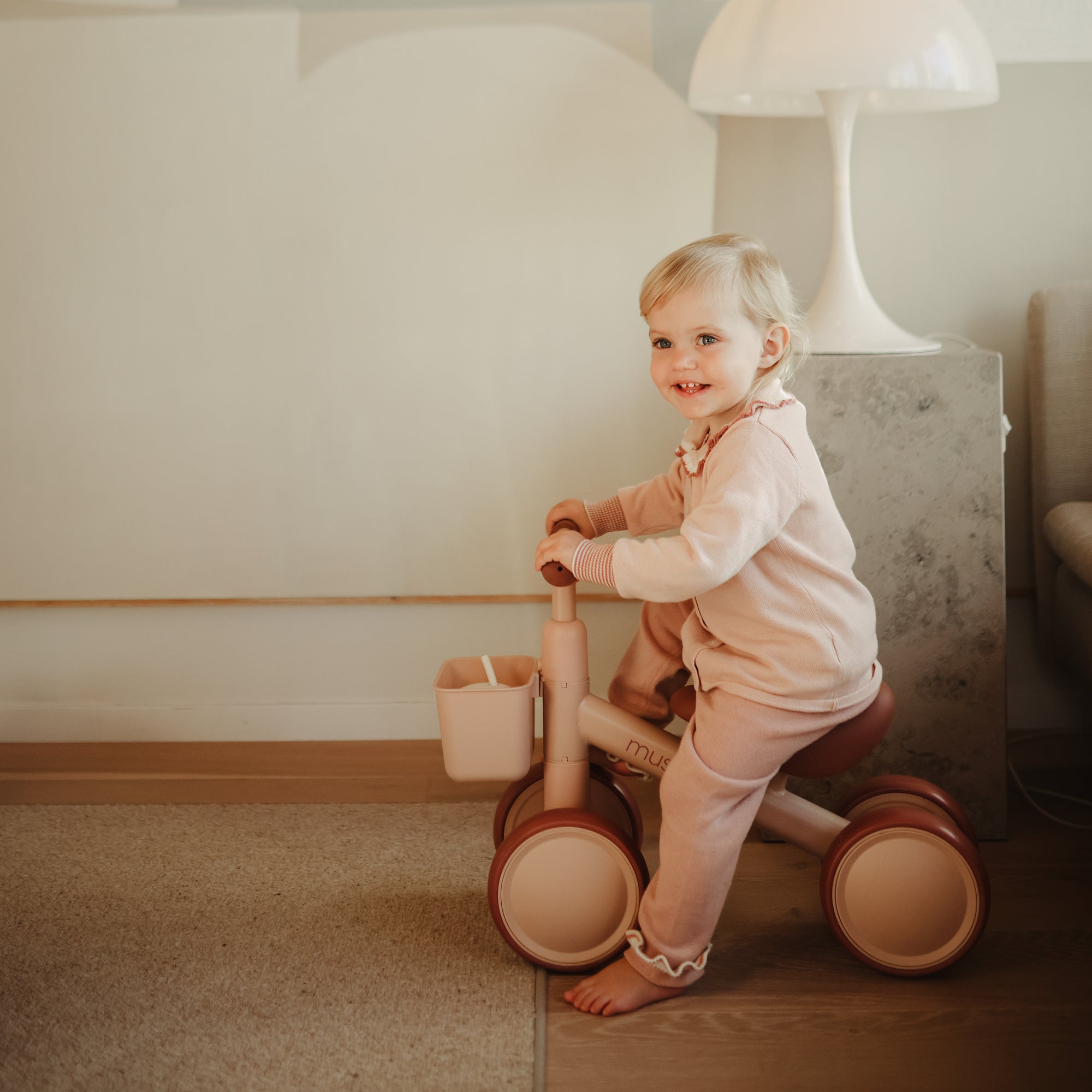 Smiling toddler in pink rides a mushie Mini Toddler Bike tricycle indoors.