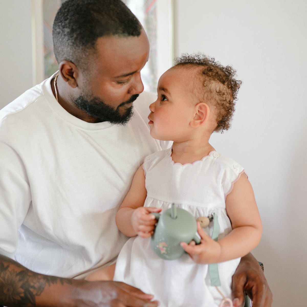 A man and toddler smile indoors while using the Mushie Silicone Training Cup + Straw.