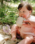 A baby in pink uses the Mushie Silicone Training Cup + Straw on a blanket near purple flowers.
