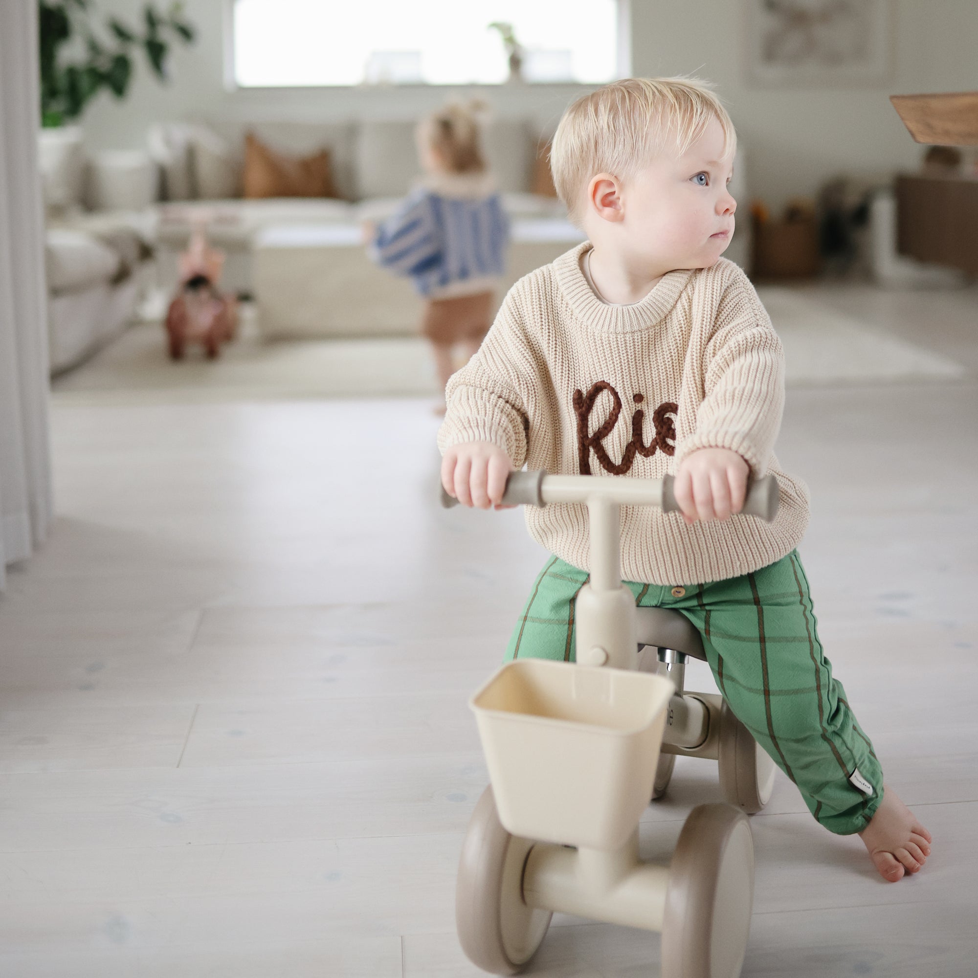 A toddler rides a beige mushie Mini Toddler Bike indoors; another child plays in the background.