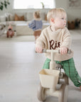 A toddler rides a beige mushie Mini Toddler Bike indoors; another child plays in the background.
