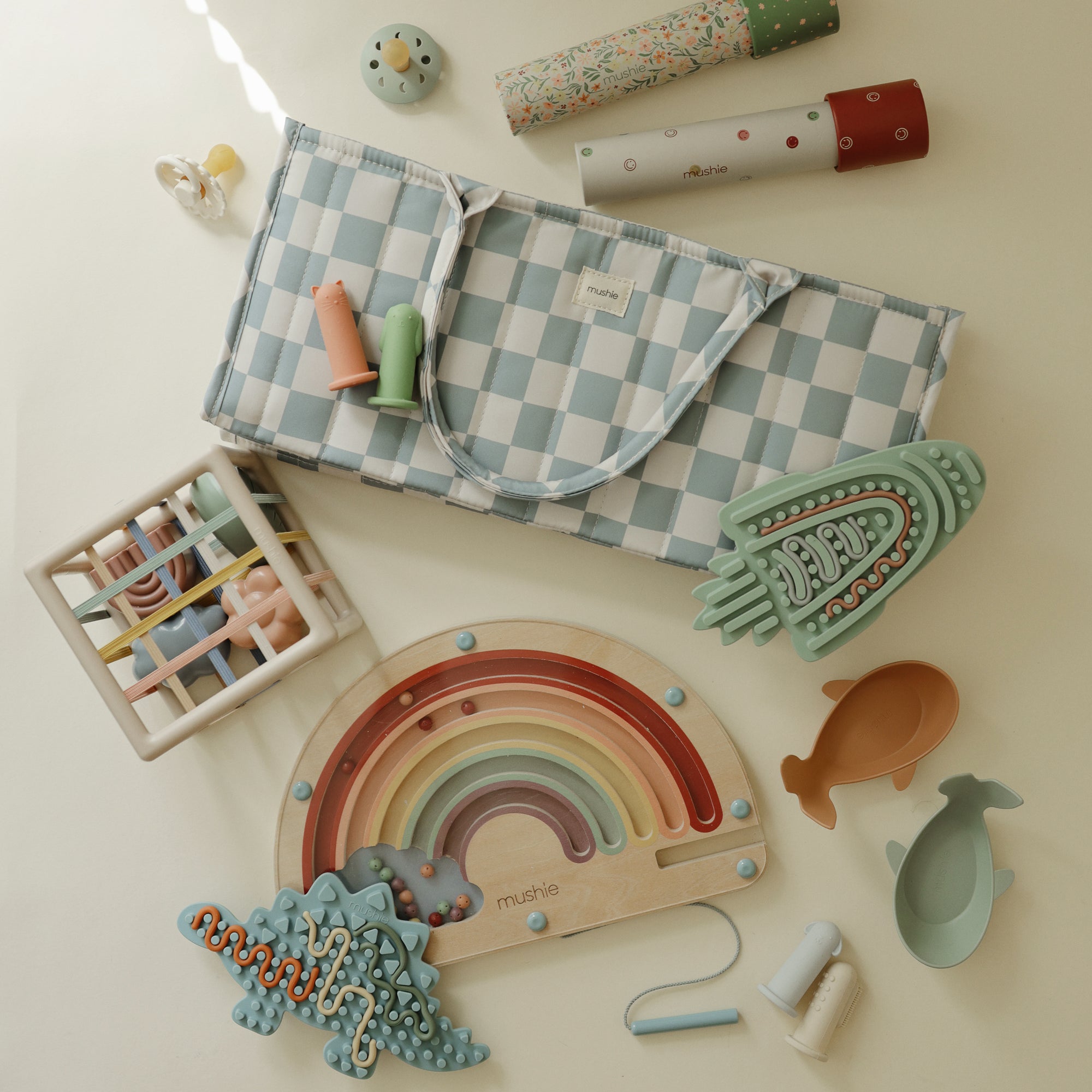 A soft play mat with pastel toys, a rattle, rainbow puzzle, and mushie Diaper Caddy nearby.