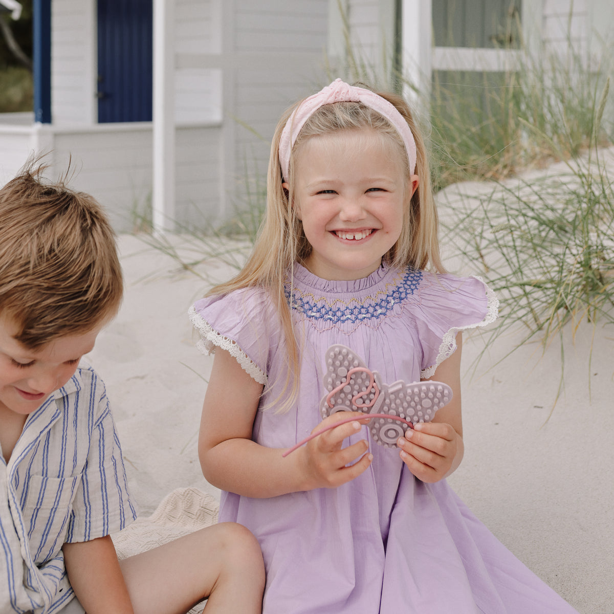Girl in purple dress holds mushie Silicone Sensory Toy sitting on sand next to a boy.