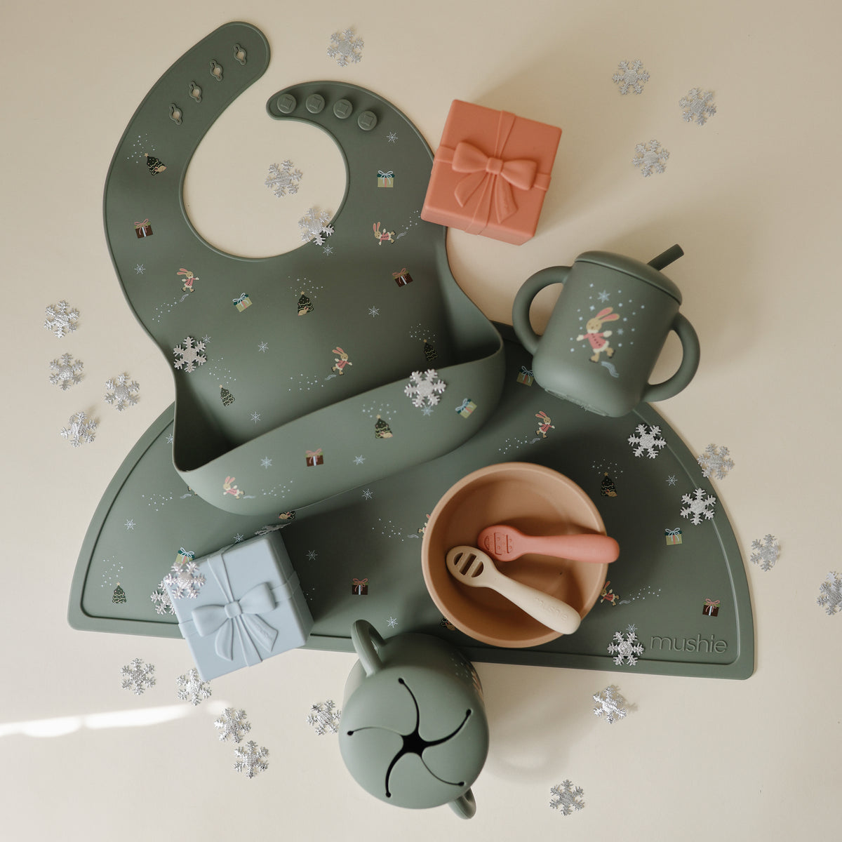 Mushie Silicone Baby Bib in sage green, perfect holiday gift with cups and snowflake confetti.