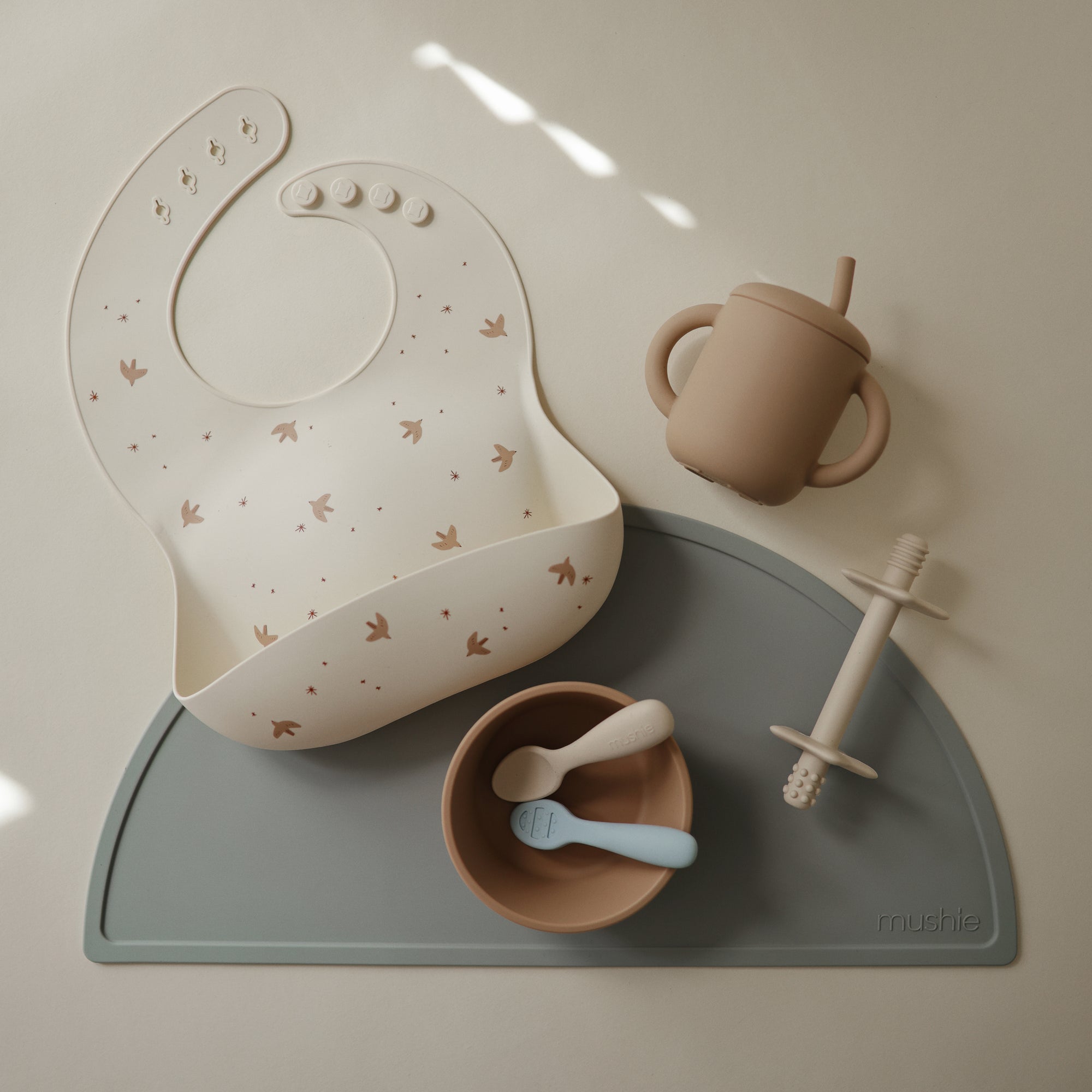 Mushie Silicone Baby Bib set: bib, cup, bowl, and teether on gray mat with adjustable neck strap.