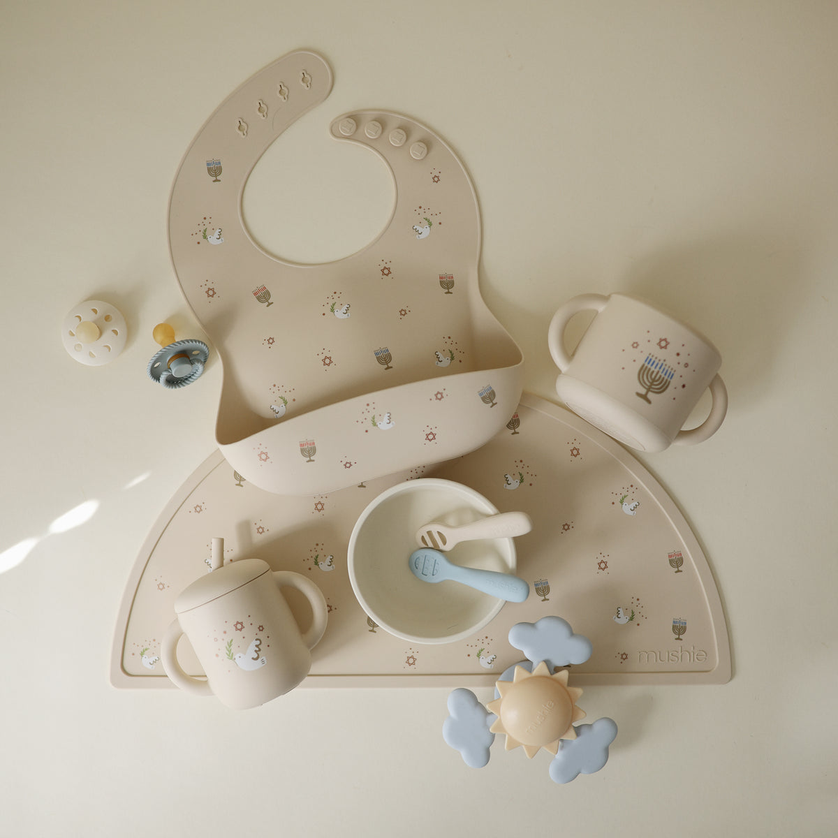 Mushie Silicone Baby Bib on a matching placemat with cups, bowl, utensils &amp; teether.