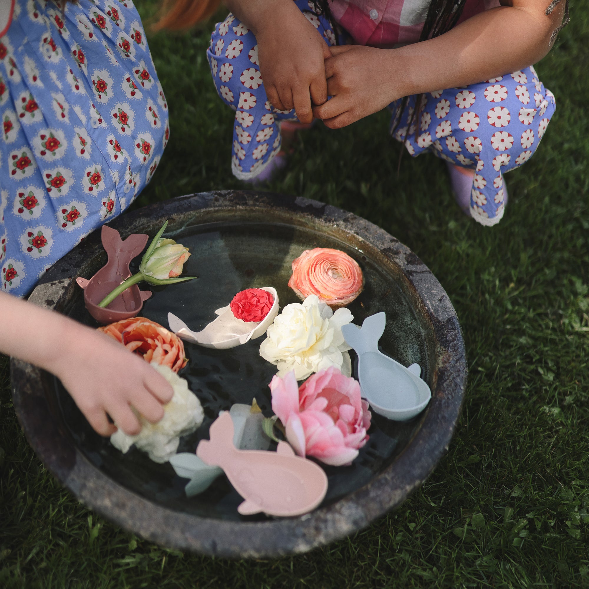 Two kids play with Mushie Whale Bath Stackers in a round water bowl on the grass.