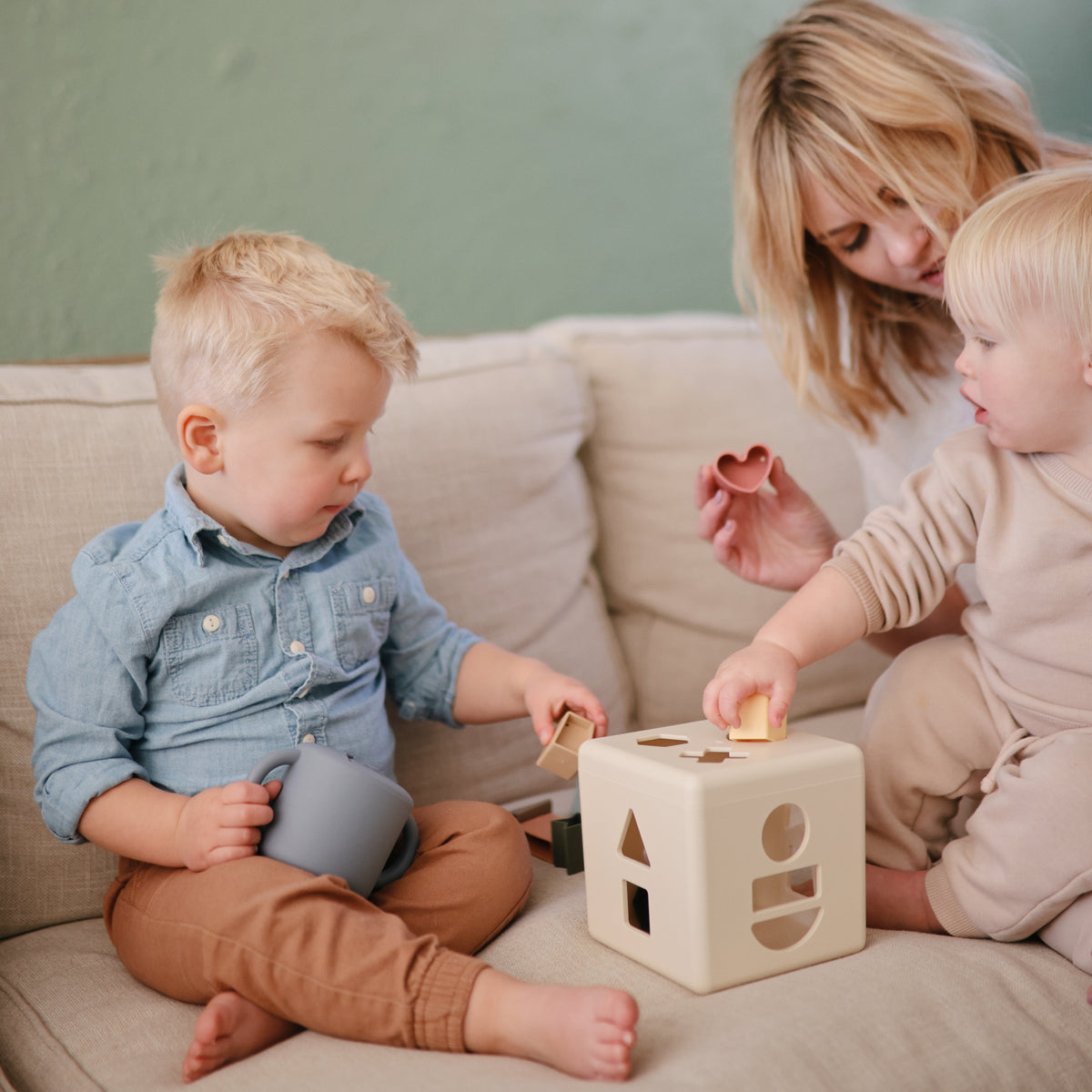 Two toddlers with a mushie Snack Cup play as an adult helps them on a beige couch.