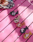 Four Mushie Whale Bath Stackers sit on a pink table under dappled sunlight.