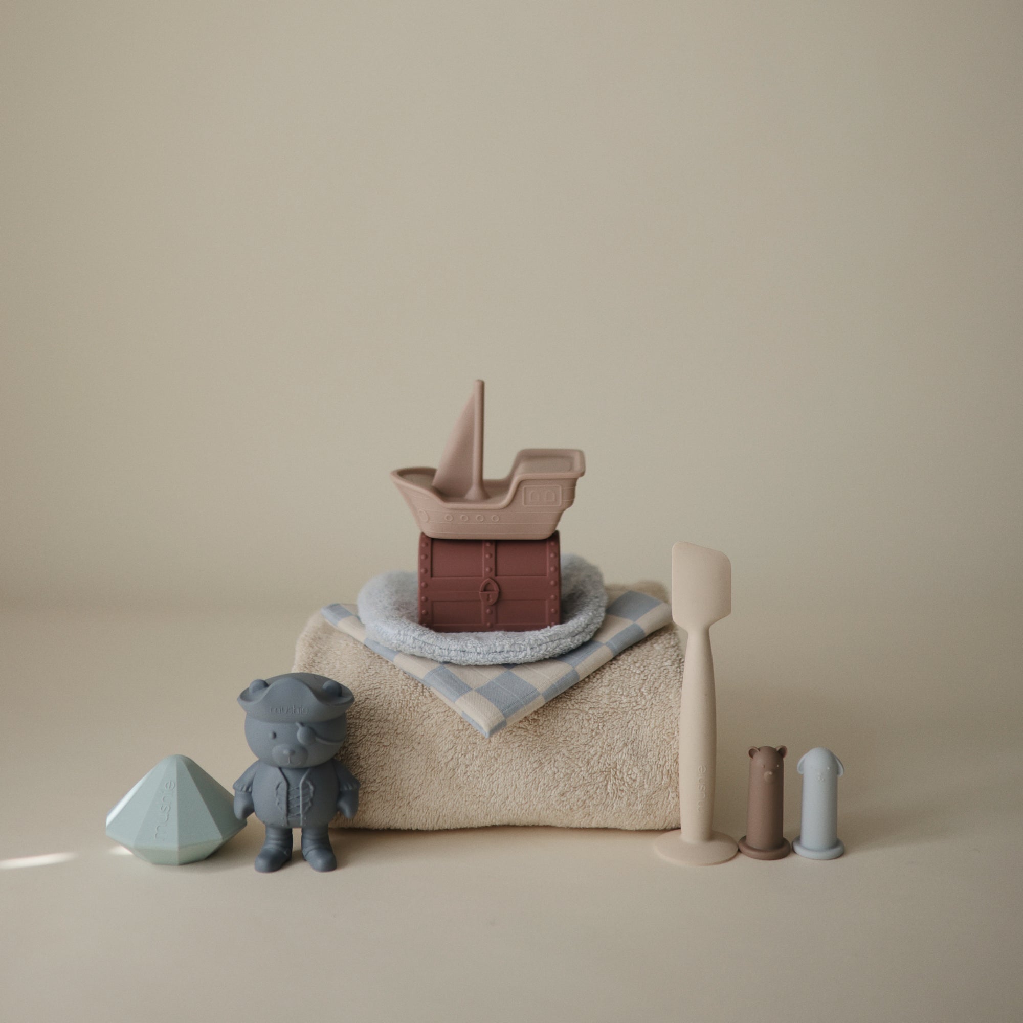 Lifestyle image of mushie bath items, including a cotton Fog hooded towel, the silicone Pirate Bath Play Set, a silicone Shifting Sand diaper applicator and silicone bear and dog Animal Finger Puppets on a neutral background. 