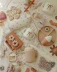Mushie Silicone Sensory Toy with pastel baby cups and plates on a cream-colored surface.