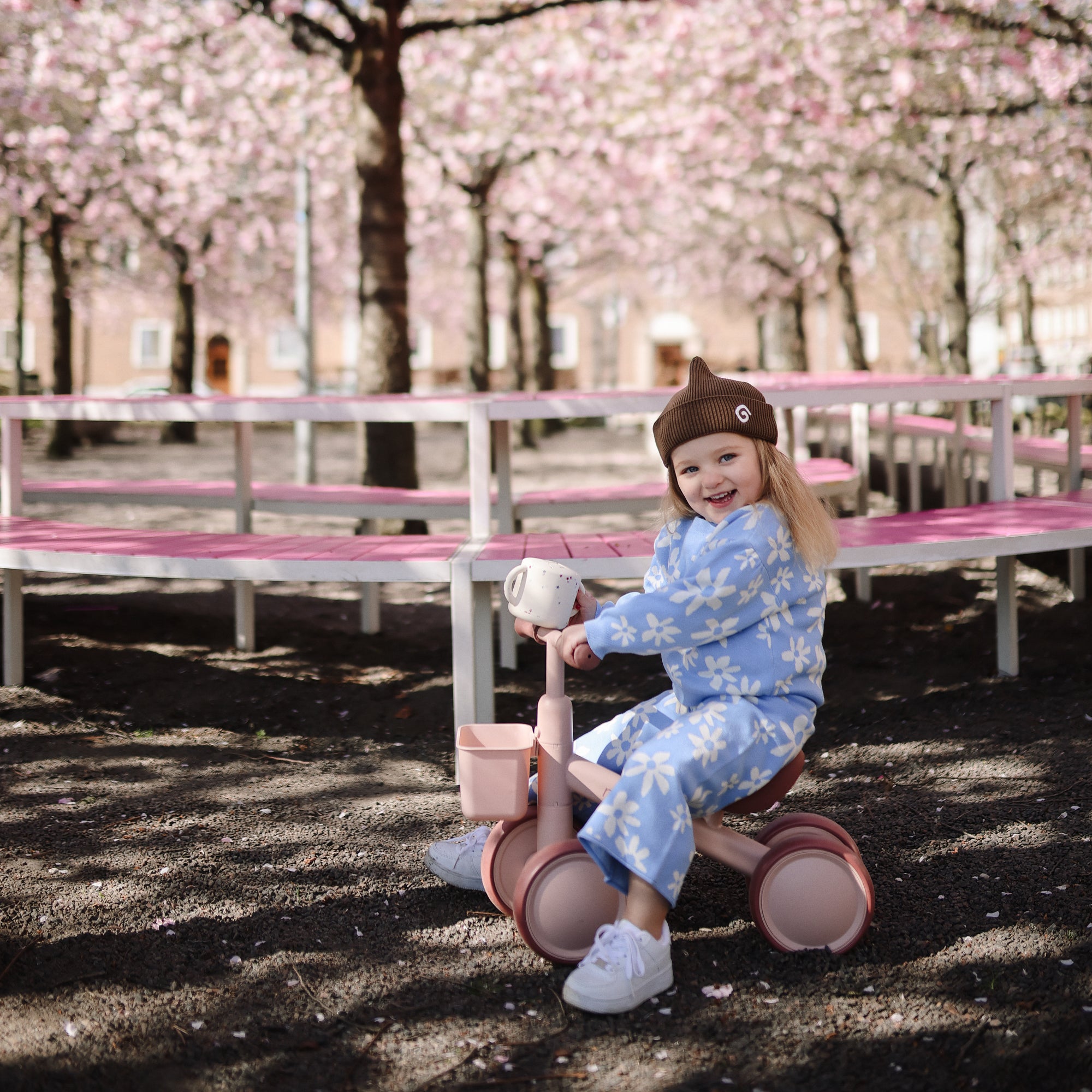 Toddler in blue floral outfit rides tricycle, holding a mushie Snack Cup.