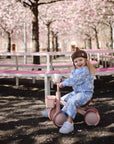 Toddler in blue floral outfit rides tricycle, holding a mushie Snack Cup.