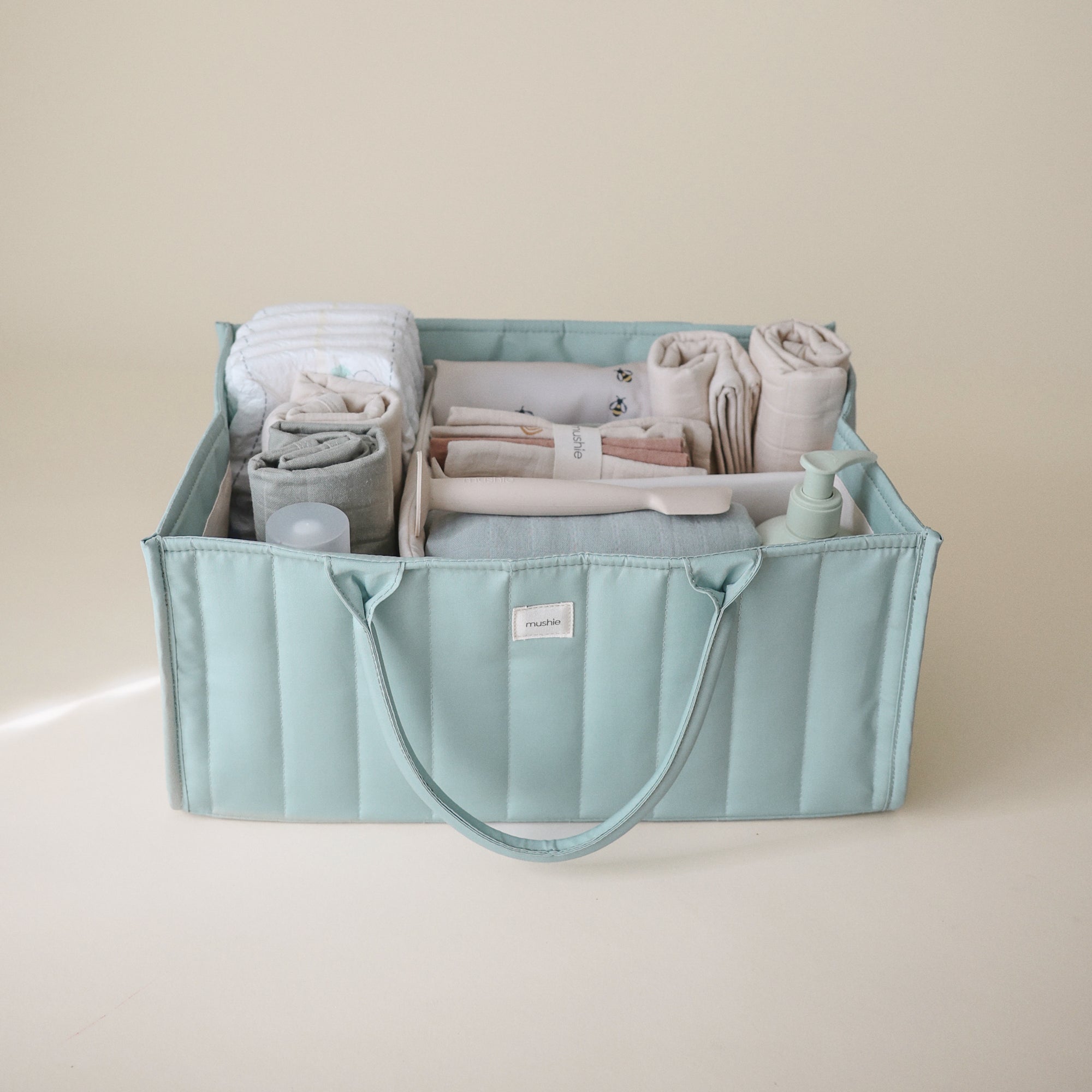 Lifestyle image of the Roman Green Diaper Caddy with folded clothes and baby nursery items on a beige background. 
