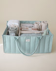Lifestyle image of the Roman Green Diaper Caddy with folded clothes and baby nursery items on a beige background. 