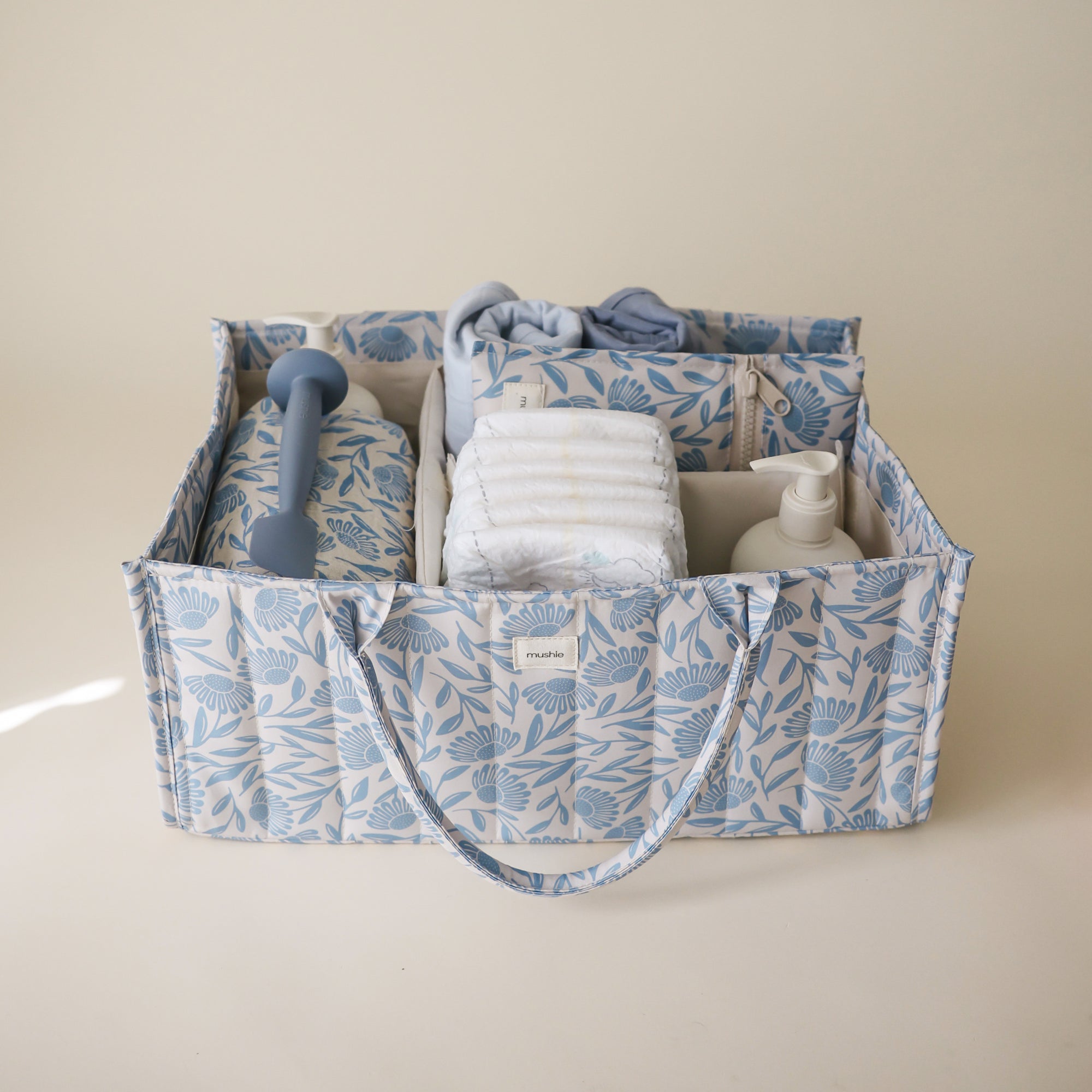 Lifestyle image of the Blue Flowers Diaper Caddy with folded clothes and baby nursery items on a beige background. 