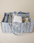 Lifestyle image of the Blue Flowers Diaper Caddy with folded clothes and baby nursery items on a beige background. 