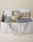 Lifestyle image of the Ivory Diaper Caddy with folded clothes and baby nursery items on a beige background. 