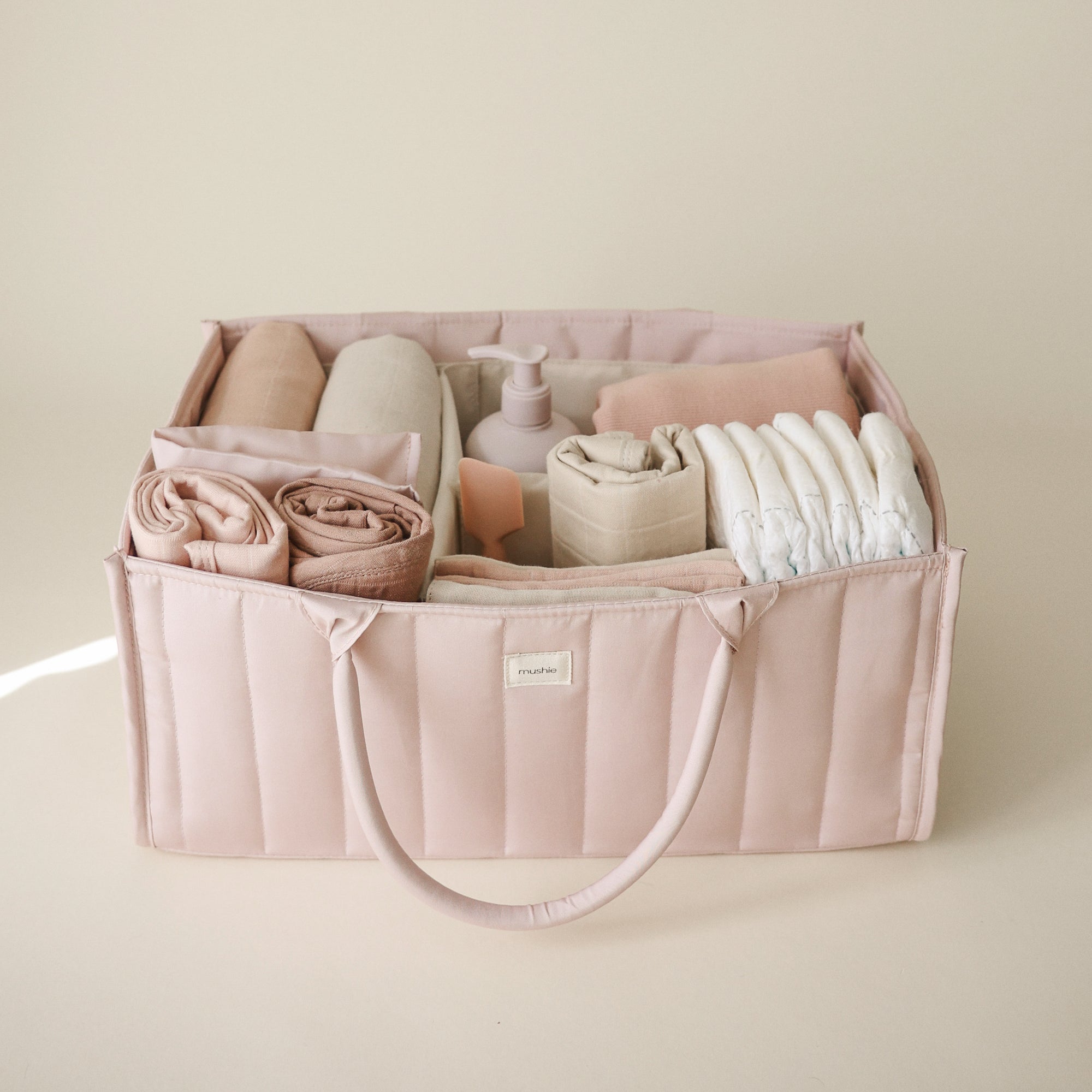 Lifestyle image of the Blush Diaper Caddy with folded clothes and baby nursery items on a beige background. 