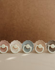 Five FRIGG Daisy Night Silicone Pacifiers in pastel pink, cream, blue, beige, and gray are lined up on a light surface with a brown background. Each pacifier features scalloped edges and comes in a 2-pack from FRIGG.