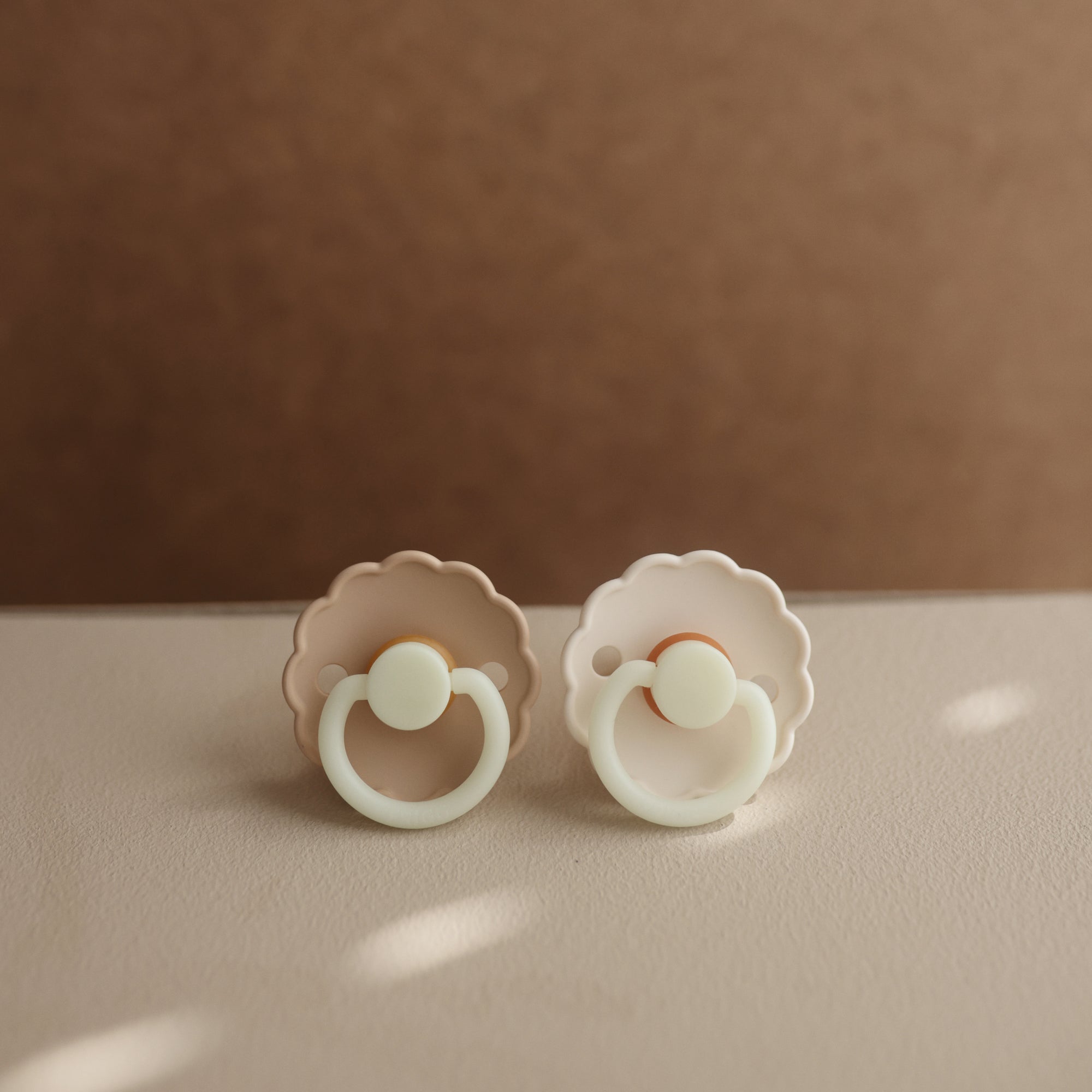 The FRIGG Daisy Night Silicone Pacifier 2-Pack features two pastel pacifiers with scalloped edges and round handles, displayed on a light surface against a soft brown background.