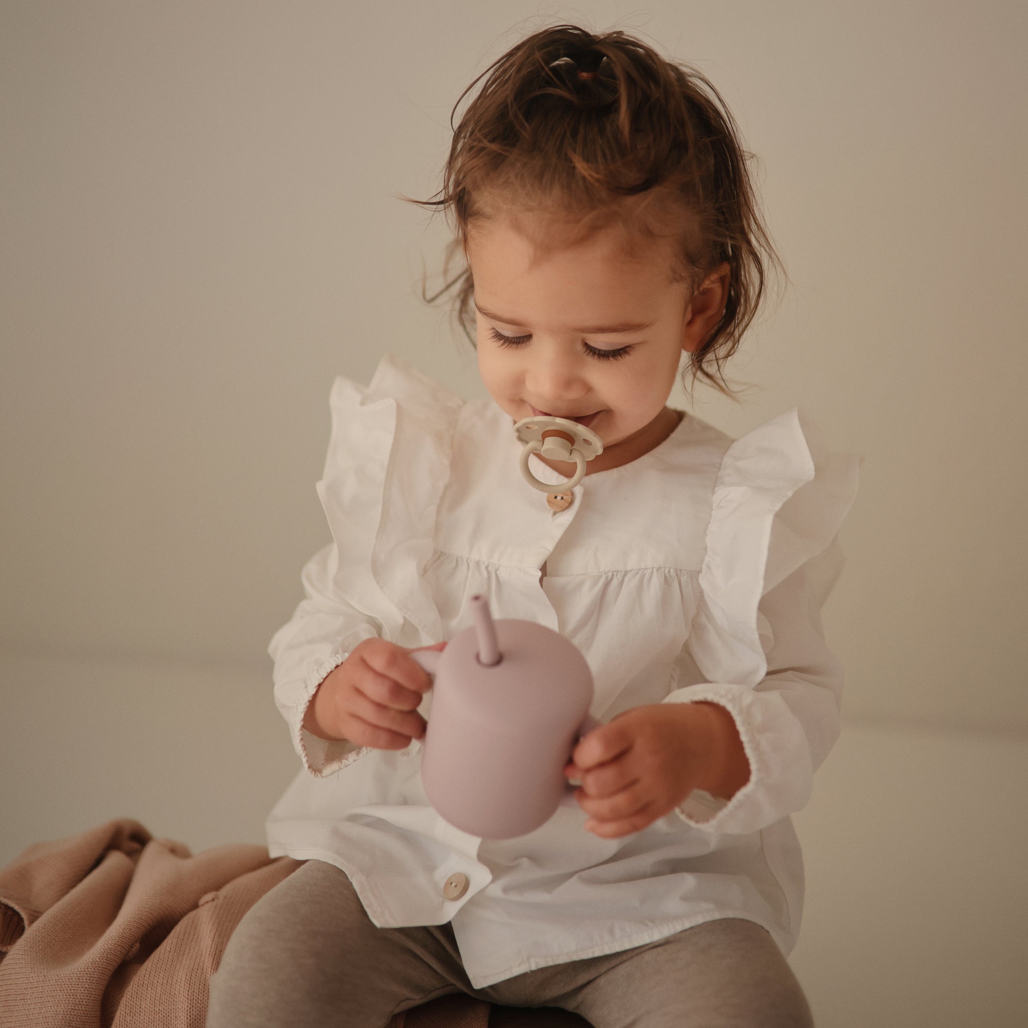 A toddler with a FRIGG Daisy Natural Rubber Pacifier holds a mauve sippy cup in white ruffles.