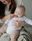 A smiling woman swaddles her baby in a mushie Organic Cotton Muslin Swaddle Blanket.