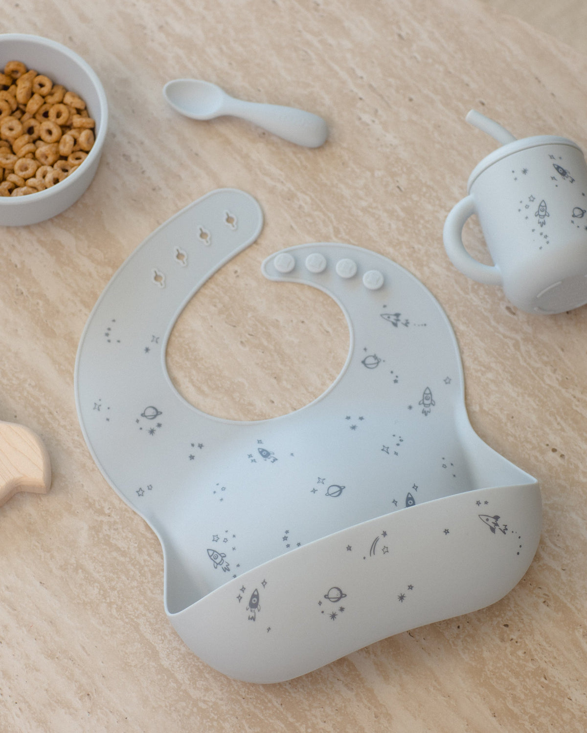 Rylee + Cru Feeding Kit Space: light blue silicone bib by cereal bowl, training cup, and spoon.