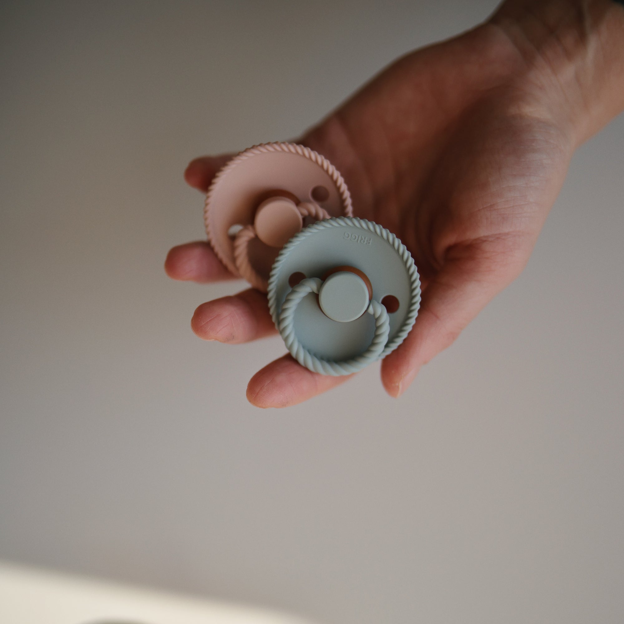 A hand holds two FRIGG Rope Natural Rubber Pacifiers by Mushie—one pastel pink, one pastel blue—both with textured edges and natural rubber latex nipples; a perfect baby essential 2-pack.