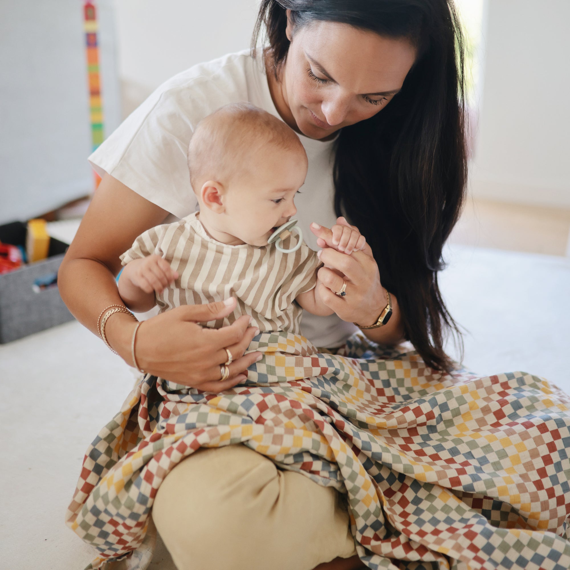A woman sits on the floor holding a baby in a striped shirt with a FRIGG Little Viking Natural Rubber Pacifier. Both are partially covered by a colorful blanket as she lovingly looks at the baby's hand.