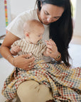 A woman sits on the floor holding a baby in a striped shirt with a FRIGG Little Viking Natural Rubber Pacifier. Both are partially covered by a colorful blanket as she lovingly looks at the baby's hand.