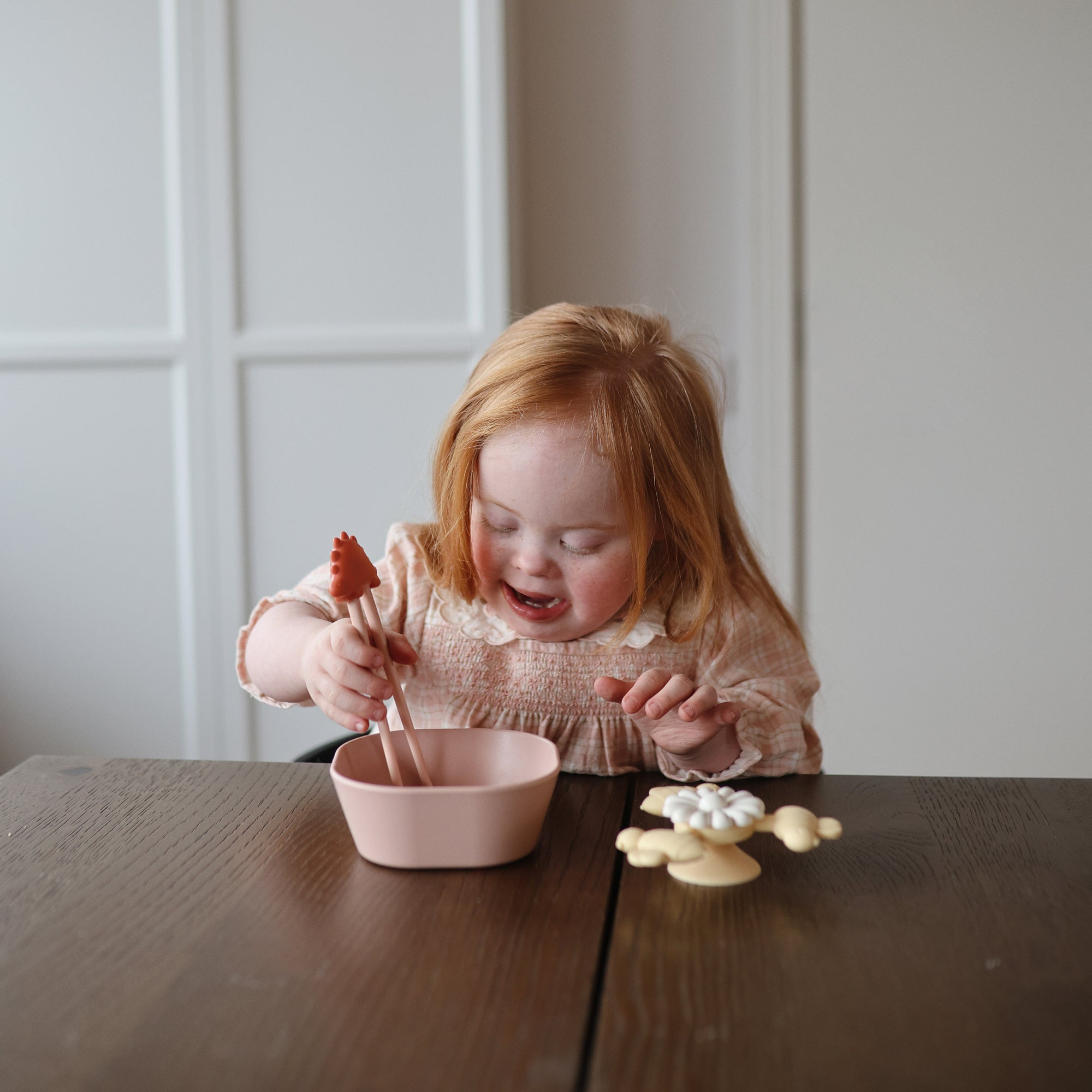 A young girl uses mushie Training Chopsticks to eat from a pink bowl at the table.