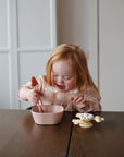 Young girl uses tongs to pick up food from a pink mushie Square Dinnerware Bowl at a table.