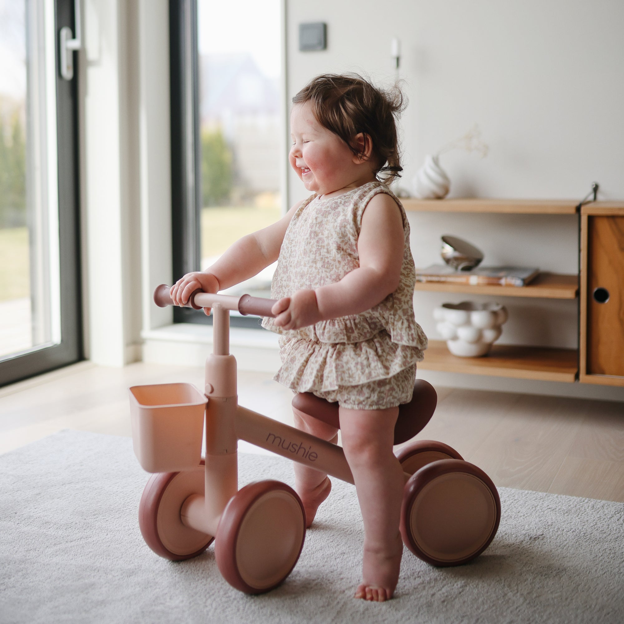 Smiling toddler rides a pink mushie Mini Toddler Bike indoors by a bright window.