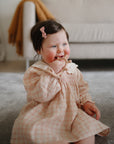 Baby in a plaid dress sits on a carpet, chewing on a mushie No-Drop Sensory Teether.